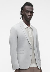 Giacca da completo grigio chiaro con rever a lancia, due bottoni e vestibilità sartoriale, indossata sopra una polo beige strutturata.