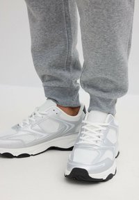 Persona con zapatillas blancas y plateadas de malla con cordones blancos y pantalones deportivos grises, de pie sobre un suelo claro.