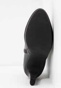 Bottine en cuir noir posée sur sa semelle, montrant le dessous lisse légèrement texturé et le petit talon carré.