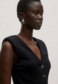 Schwarzes ärmelloses Kleid mit V-Ausschnitt, drei Knöpfen vorne und einer kleinen Tasche. Strukturierter Stoff mit tailliertem Schnitt. Silberne Ohrringe.