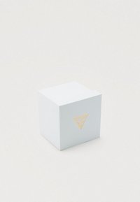 Bílá krabice ve tvaru krychle se zlatým logem Guess uprostřed jedné strany, umístěná na jednoduchém světlém pozadí.