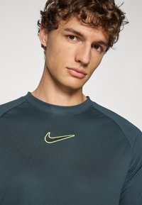 Nike sporta krekls tumši teal krāsā, ar melnu swosh logo, ko apvilkusi neona dzeltena apmale, izgatavots no gludas, pieguļošas auduma ar apaļu kakla izgriezumu.