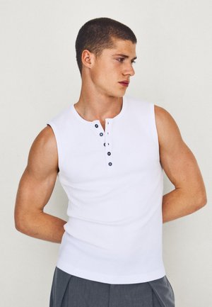 Giovane uomo con i capelli corti che indossa una camicia bianca senza maniche con bottoni e pantaloni grigi, guarda verso destra con le mani dietro la schiena.