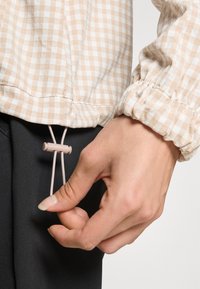 Beige and white checked fabric with elastic cuffs, featuring a drawstring with a toggle for adjustment, paired with dark pants.