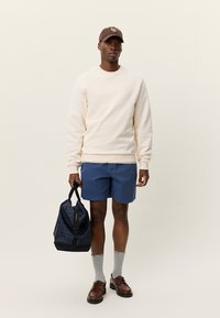 Κρεμ φούτερ, μπλε σορτς, γκρι κάλτσες, καφέ παπούτσια και μια σκοτεινή τσάντα με καρό σχέδιο. Το καπέλο έχει λογότυπο. Απλή, καθημερινή εμφάνιση.