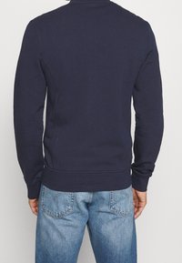 Persona che indossa un maglione a maniche lunghe blu navy e jeans di denim azzurri, in piedi con la schiena rivolta verso la telecamera.