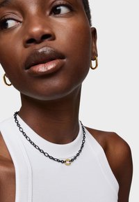 Collana a catena nera con un anello d'accento dorato, indossata su una maglietta bianca a coste senza maniche. Sono visibili anche orecchini a cerchio dorati.