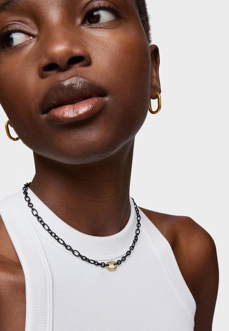 Collana a catena nera con un anello d'accento dorato, indossata su una maglietta bianca a coste senza maniche. Sono visibili anche orecchini a cerchio dorati.