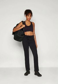 Frau im schwarzen Sport-BH und -Hose, die eine große schwarze Sporttasche über der Schulter hält, vor einem schlichten hellen Hintergrund stehend.