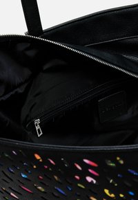 Black tote bag with perforated design revealing colorful interior. Features a zip pocket and smooth leather material. Lined with black fabric.