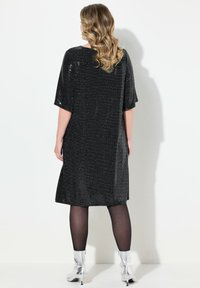 Vestido negro brillante con mangas cortas, corte suelto, hasta la rodilla, combinado con medias transparentes y botines metálicos. Diseño trasero minimalista.