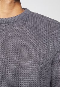 Zbliżenie na osobę w szarym swetrze z fakturowanym splotem, z prążkowanym dekoltem okrągłym na tle jednolitym.