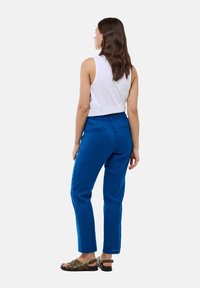 Pantalon bleu en tissu lisse, avec une coupe décontractée et des poches latérales. Porté avec un top sans manches blanc et des sandales.