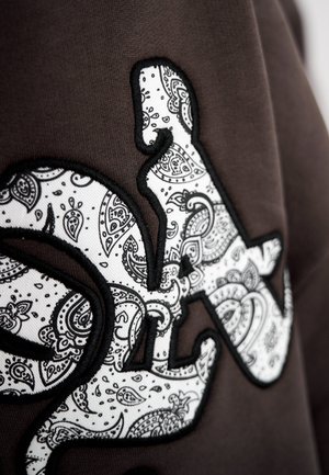 Brown fabric featuring a textured design with a white paisley pattern outlined in black, forming the shape of stylized lettering.