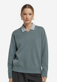 Helles blau-grünes, übergroßes Sweatshirt mit V-Ausschnitt, geripptem Saum und langen Ärmeln, über einem blauen Hemd mit Kragen getragen. Weiches, strukturiertes Material.