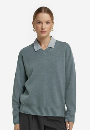 Un pull surdimensionné bleu-vert clair avec un col en V, un ourlet côtelé et des manches longues, superposé sur une chemise à col bleue. Tissu doux et texturé.