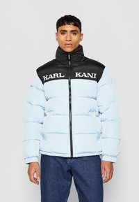 Lyseblå pufferjakke med svart krage, glidelås foran og hvit merkevarelogo "KARL KANI" over brystet. Har sydd paneler.
