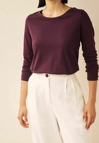 Langärmliges, rundhalsiges Oberteil aus tiefviolettem Baumwollstoff, kombiniert mit hoch taillierten, cremefarbenen Leinenhosen mit Knopfdetail.
