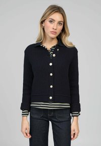 Cardigan en maille bleu marine avec texture côtelée, fermeture à boutons et poignets rabattus, porté sur une chemise rayée foncée. Jean en denim complète la tenue.
