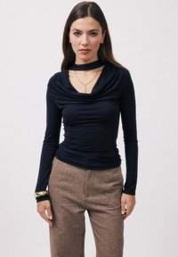 Haut noir à manches longues avec un col bénitier drapé, tissu doux et coupe ajustée, associé à un pantalon taille haute beige et un bracelet doré.