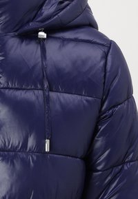 Marineblå puffjakke med glanset finish, som har hette, quiltet design og en sølvfarget snorjustering.