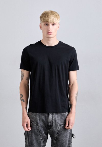 Jeune homme aux cheveux blonds, portant un t-shirt noir uni et un jean gris, se tenant contre un fond gris clair uni.
