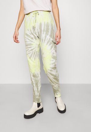 Person iført lysegrønne og grå tie-dye joggingbukser med sidelomme, hvid elastisk taljebånd og hvide ankelstøvler med sorte såler.