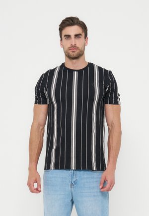 Mand iført sort kortærmet skjorte med lodrette hvide striber og lyseblå jeans, stående foran en ensfarvet hvid baggrund.