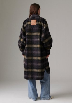 Femme portant un long manteau à carreaux foncé avec une pièce beige sur le haut du dos, associé à un jean large bleu et des chaussures claires.