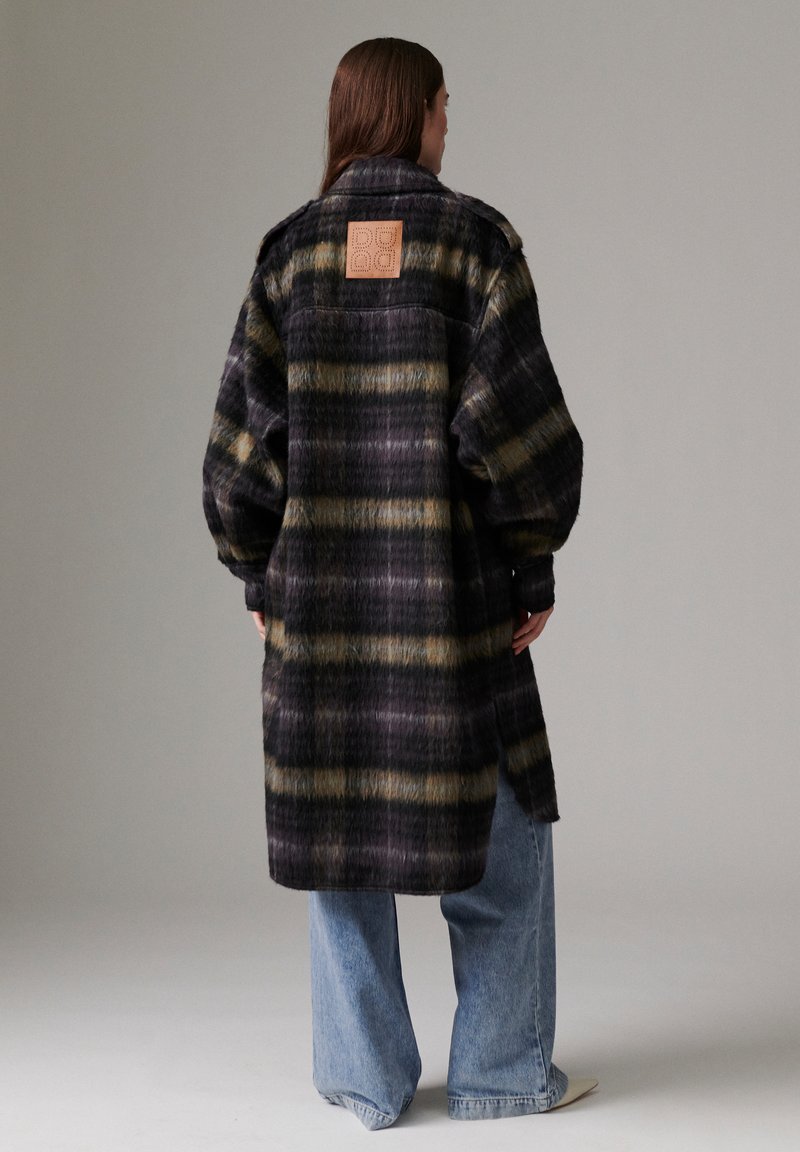 Femme portant un long manteau à carreaux foncé avec une pièce beige sur le haut du dos, associé à un jean large bleu et des chaussures claires.