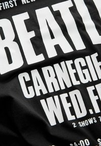 Schwarzes T-Shirt mit großen weißen Druckbuchstaben, die eine Show bewerben. Der Text enthält "BEATLES", "CARNEGIE", "MITTWOCH, FREITAG" und Ticketpreise, mit einer weichen Baumwolltextur.