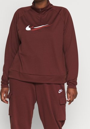 Person iført vinrød Nike træningsdragt med lagdelt swoosh-logo på toppen og hvidt Nike-logo på lommen af cargobukserne.