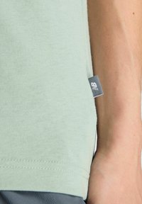 Light green short-sleeve shirt made of soft fabric. Features a side tag with logo, showing stitching detail at the hem and sleeve.