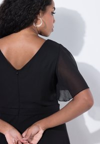 Femme aux longs cheveux bouclés portant des boucles d'oreilles créoles et une robe noire à manches transparentes, vue de dos contre un mur blanc.