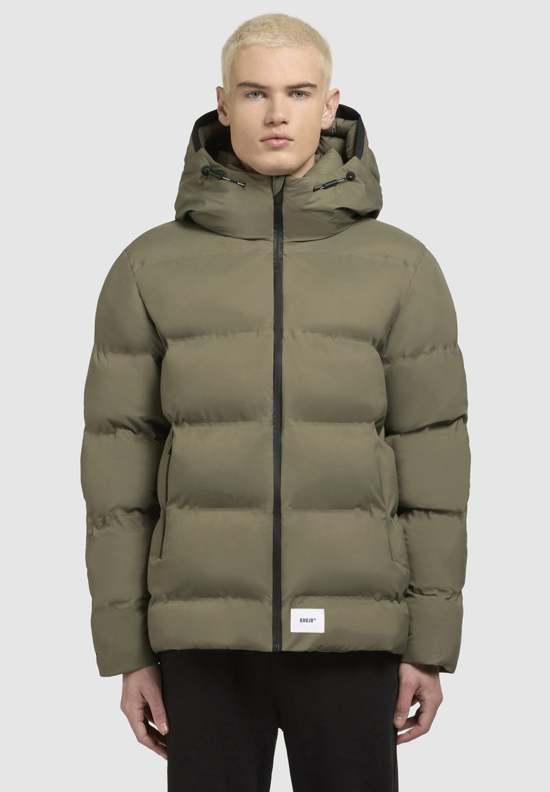 Chaqueta de plumas verde oliva con capucha, cremallera negra y parche con logo. Presenta un diseño acolchado y bolsillos laterales. Textura suave y mate.
