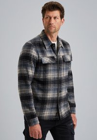 Mies, joka on pukeutunut sinivalko-mustaruutuiseen napitettavaan paitaan, jossa on rintataskut, ja mustiin housuihin, katsoo vasemmalle yksinkertaista taustaa vasten.