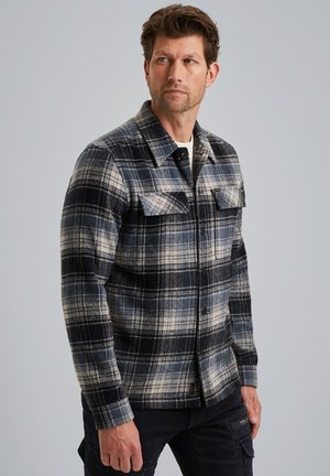 Ein Mann trägt ein kariertes Button-Up-Hemd in Blau, Schwarz und Weiß mit Brusttaschen sowie schwarze Hosen und schaut nach links vor einem schlichten Hintergrund.