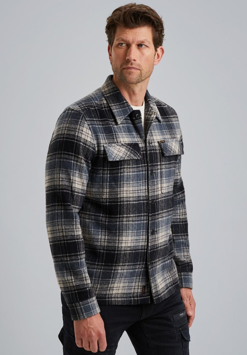 Mies, joka on pukeutunut sinivalko-mustaruutuiseen napitettavaan paitaan, jossa on rintataskut, ja mustiin housuihin, katsoo vasemmalle yksinkertaista taustaa vasten.