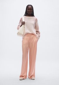 Pull rose clair en maille avec manches larges, associé à un pantalon ample couleur pêche douce. Porte un sac blanc et des ballerines beiges.