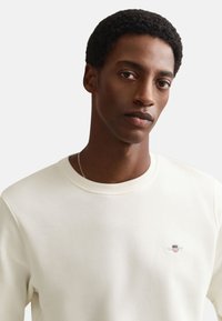 Νέος άνδρας με κοντά σγουρά μαλλιά που φοράει λευκό φούτερ με στρογγυλή λαιμόκοψη και μικρό λογότυπο στο στήθος, με φόντο σε απλό χρώμα.