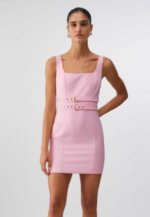 Donna che indossa un mini abito rosa senza maniche con scollo quadrato e due cinture abbinate in vita, in piedi contro uno sfondo semplice.