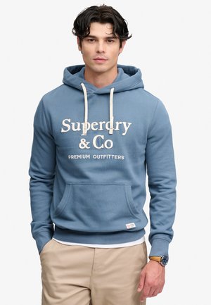 Ung mand iført en blå Superdry & Co hættetrøje med hvide snore og beige bukser, stående med hænderne i lommerne.