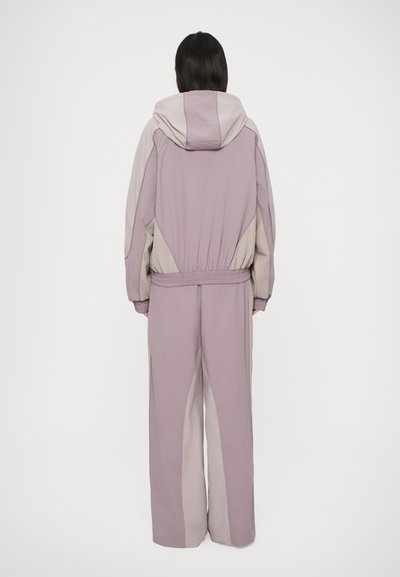 Άτομο με την πλάτη στραμμένη, φορώντας ένα δίχρωμο μωβ και μπεζ φούτερ με κουκούλα, με ελαστικούς ιμάντες και παντελόνι φαρδύ, μπροστά σε απλό λευκό φόντο.