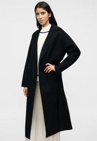 Μαύρο μακρύ παλτό με φαρδιά γιακά, ανοιχτό μπροστά και άνετη εφαρμογή. Φορεμένο πάνω από ένα λευκό πουλόβερ, με λεία υφή και μίνιμαλ σχεδίαση.