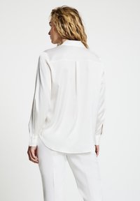 Donna con capelli biondi ondulati che indossa una camicetta bianca a maniche lunghe e pantaloni bianchi, vista di spalle su uno sfondo chiaro semplice.