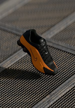 Basket Nike noire et orange avec une semelle visible à coussin d'air flottant au-dessus d'une surface en grille métallique texturée.