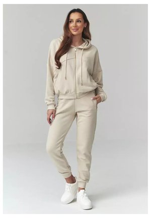 Kobieta stojąca w beżowej bluzie z kapturem na zamek i pasujących spodniach dresowych oraz białych trampkach, z rękami w kieszeniach, na jednolitym jasnym tle.