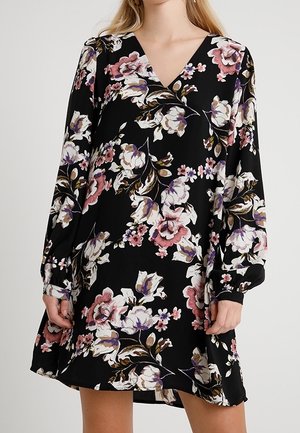 Femme portant une robe noire à manches longues avec un imprimé floral rose, blanc et violet et un décolleté en V, les mains détendues le long du corps.