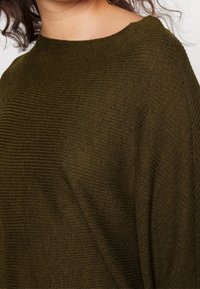 Olijfgroene gebreide trui met een geribbeld patroon, met een hoge halslijn en een losse pasvorm. De stof lijkt zacht en warm.