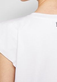 Bílá bavlněná tričko s krátkým rukávem, s kulatým výstřihem. Malý logo detail je viditelný blízko zadního výstřihu. Hladká textura.